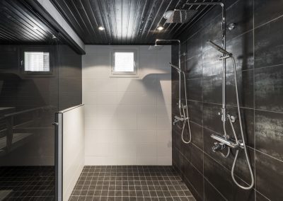 Pesuhuone kahdella suihkulla ja saunatilassa puolilasiseinä. Lattiassa tumma laatoitus. Seinät tummalla ja valkoisella laatoituksella.