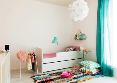 Vaalea lastenhuone, jossa värikäs matto ja turkoosin sävyiset verhot.