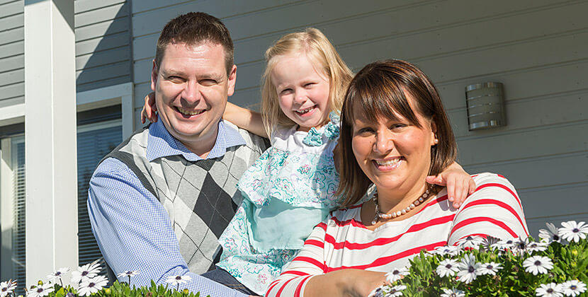 Onnellisesti hymyilevä perhe uuden dekotalon terassilla. Perheen tyttö istuu iloisena vanhempien välissä.