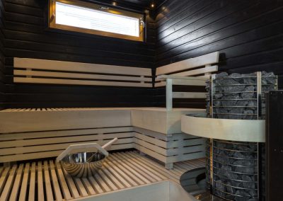 Sauna jossa mustat seinäpaneelit ja vaaleat lauteet joihin upotettu kiuas.