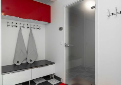 Mustavalkoinen kodinhoitohuone, jossa mustavalkoruutuinen lattialaatoitus ja punaiset kaapinovet, matto ja roskakori.
