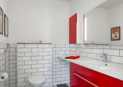 Mustavalkoinen WC, jossa mustavalkoruutuinen lattialaatoitus ja punaiset kaapinovet. WC-istuin on seinään kiinnitetty ja WC -kotelo ja huoneen seinät on laatoitettu seinän puolivälistä alaspäin valkoisella laatalla tiililadontana.